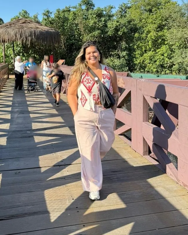 Maria Rejane sorrindo enquanto caminha por uma passarela de madeira, num dia de sol