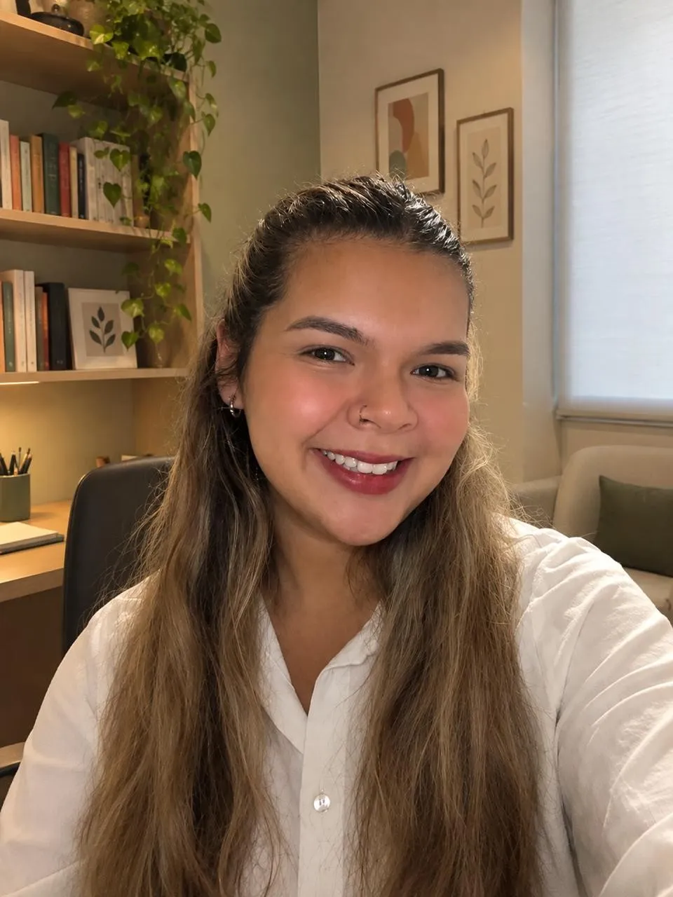 Maria Rejane sorrindo no consultório, com estante e plantas ao fundo